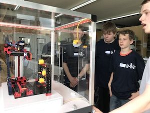 NFT-Azubi Kai Grothaus steht mit Schülern vor einem Glasschrank mit dem Modell einer Automatisierungsanlage